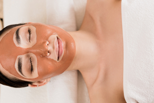 top view of beautiful woman lying with clay mask on face with closed eyes at spa  - Photo, Image