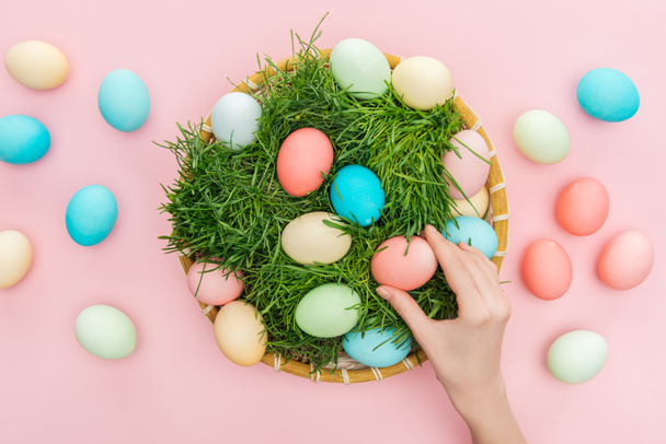 top view of female hand with easter eggs in wicker plate with grass isolated on pink  - Фото, зображення