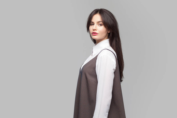 Side  view portrait of serious beautiful young woman with makeup and brunette hair in white shirt and brown dress standing and looking at camera on grey background - Φωτογραφία, εικόνα