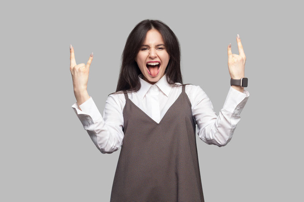 Portrait of amazed beautiful young woman with makeup and brunette hair in white shirt and brown dress showing with rock gestures and looking at camera with tongue out on grey background - Foto, afbeelding