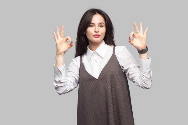 Portrait of beautiful young woman with makeup and brunette hair in white shirt and brown dress looking at camera and showing Ok sign gesture on grey background. - Foto, imagen