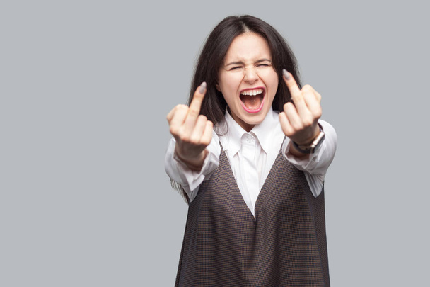 Portrait of angry young brunette hair woman in white shirt and brown dress showing with middle fingers fuck sign gestures and screaming on grey background - Photo, image