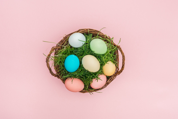 top view of easter eggs in wicker basket with grass isolated on pink - Foto, Bild