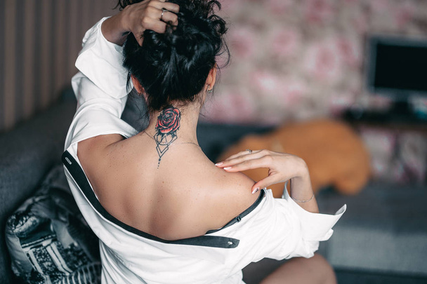Back neck tattoo of a woman brunette girl in shirt at home - Foto, imagen