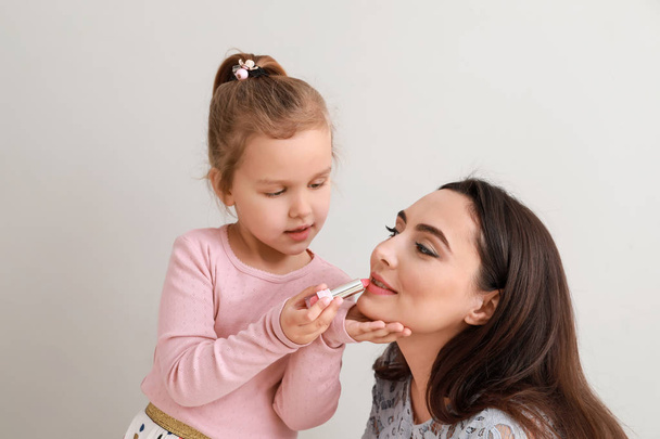 Cute little daughter applying lipstick onto mother's lips against light background - Photo, Image