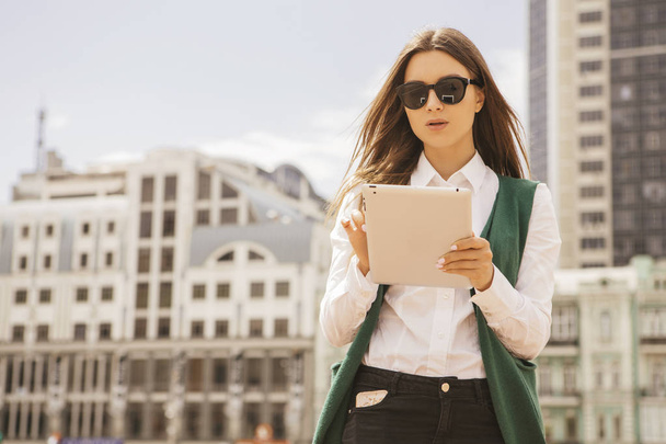 Mooie brunette zakenvrouw in witte blouse en groene jas op een tablet in haar handen buitenshuis werken. Freelancer in Europese stad. Ruimte voor tekst. - Foto, afbeelding