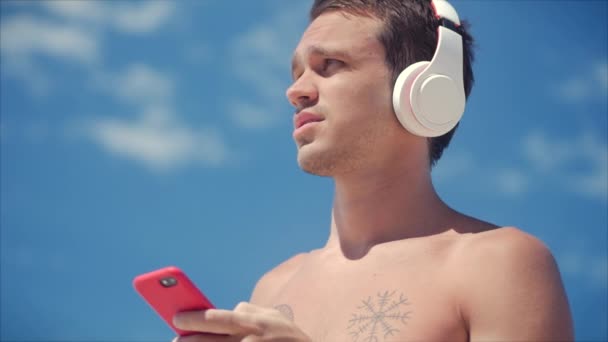 Bello Giovane Ascoltare musica dal suo smartphone in cuffie bianche senza fili, Ballando sulla spiaggia in background Blue Sky
. - Filmati, video
