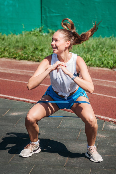 a young woman performs high jump exercises with a fitness elastic band on her feet on the sports ground - 写真・画像
