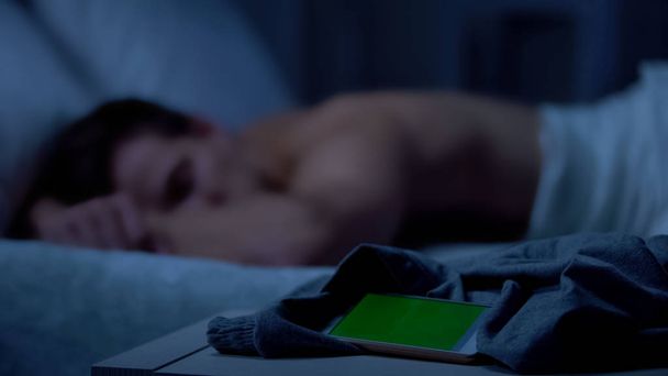 Handy mit grünem Bildschirm auf dem Bett liegend, müder Mann schlafend im Hintergrund - Foto, Bild
