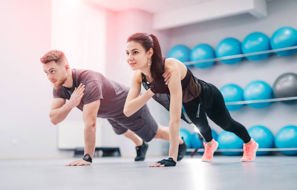 Pretty young couple doing exercises together in the light gym. Sporty male and female in sportswear training horizontal to the floor by one hand making balance - Foto, afbeelding