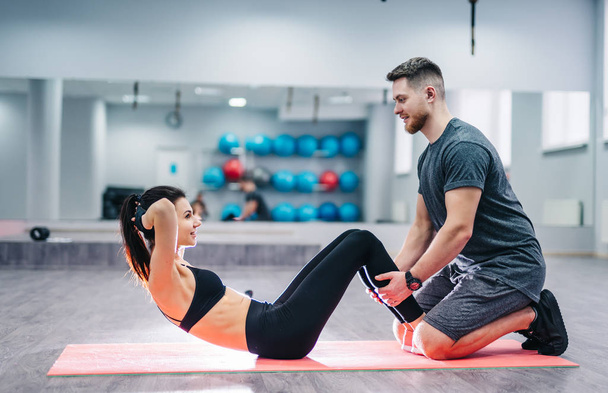 Side view of a girl doing press exercises on the mat with the help of a smiling male. Happy man and woman in a good sport shape doing exercises. - Valokuva, kuva
