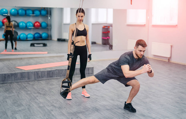 Strong beardy trainer doing exercise with TRX and a woman in sportswear looking at him in gym. Pretty female standing near handsome man doing legs training on TRX. Sport concept - Foto, Bild