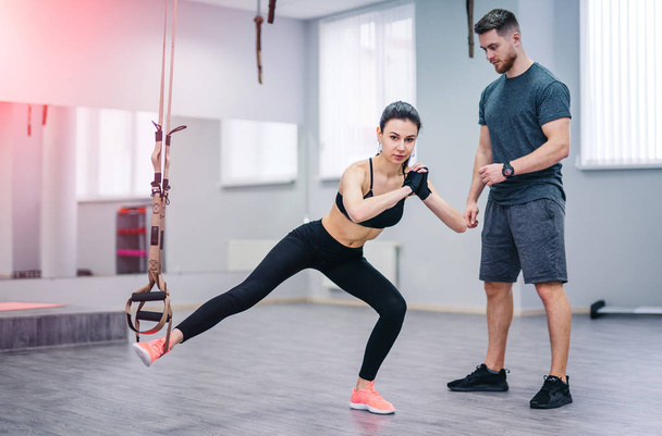 Young girl doing suspension rope exercise together with her personal coach on the gym background. Beautiful female doing legs training on TRX with private instructor indoors - Foto, afbeelding