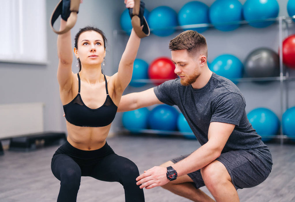 Muscular beardy trainer helping attractive female to practice with TRX in the fitness center. Sporty woman doing exercises with personal instructor at gym. Close-up - Zdjęcie, obraz