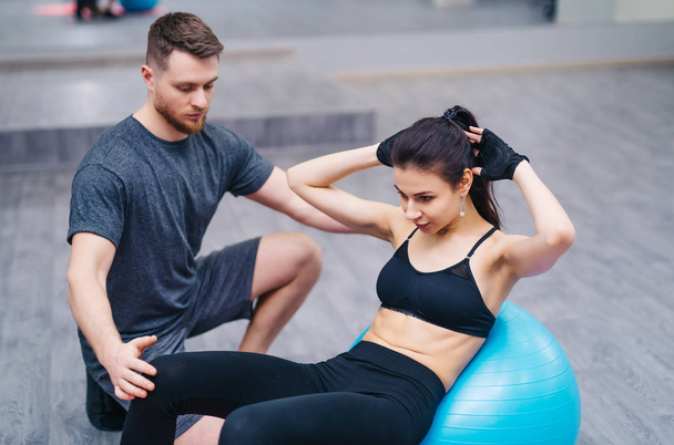 Attractive young woman doing abs on a fitness ball with personal trainer on the floor in gym. Sporty girl leans her body sitting on a ball with arms behind her head and a strong male coach - Photo, image