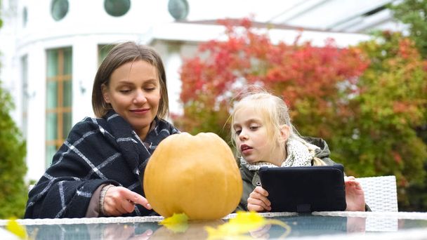 Mother carefully carving pumpkin while daughter looking for tips in internet - Foto, Imagem