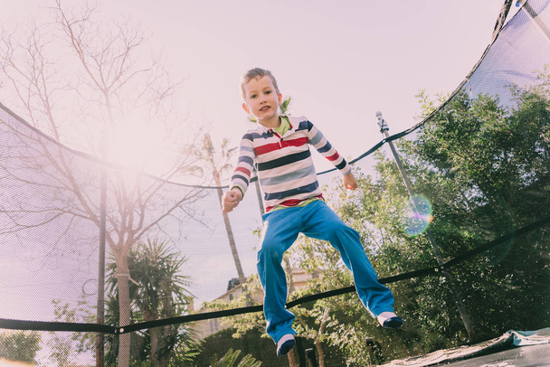 5 χρονών αγόρι άλμα σε ένα τραμπολίνο άσκηση στην πίσω αυλή του σπιτιού του, απολαμβάνοντας την άνοιξη με τη χειρονομία της ευτυχίας και της ζωής. - Φωτογραφία, εικόνα