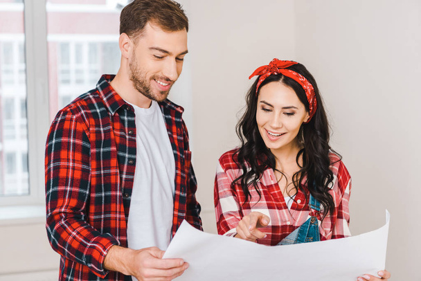 cheerful woman pointing with finger at blueprint while standing with boyfriend at home - Foto, Imagem