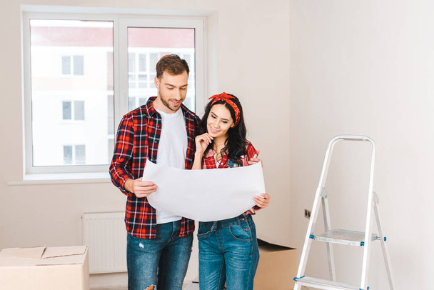 cheerful couple  standing near boxes and holding blueprint at home - Foto, Imagem
