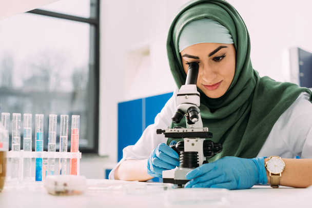 beautiful female muslim scientist looking through microscope during experiment in chemical laboratory - Photo, Image