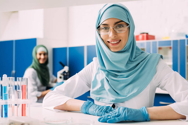 χαμογελαστός, γυναίκα μουσουλμάνος επιστήμονας με σωλήνες δοκιμής που βλέπουν τα φωτογραφικών μηχανών κατά τη διάρκεια του πειράματος σε χημικό εργαστήριο  - Φωτογραφία, εικόνα