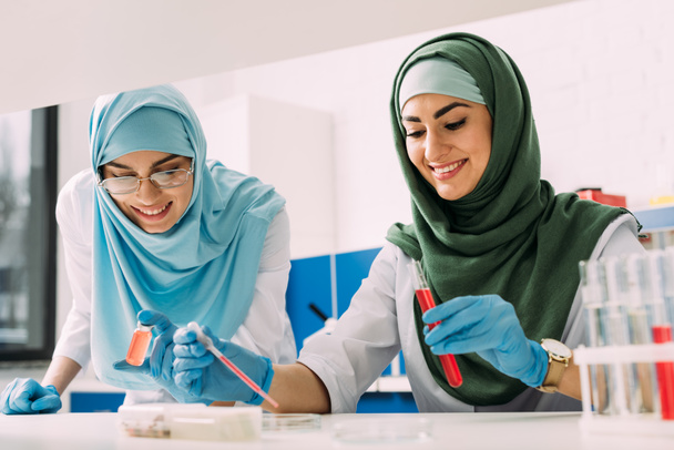 científicos musulmanes sonrientes en hijab con tubo de ensayo y pipeta durante el experimento en laboratorio químico
 - Foto, Imagen
