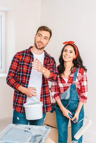 cheerful couple holding paint bucket and roller while talking at home - Zdjęcie, obraz