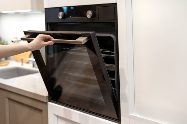 Женщина открывает дверь новой современной встроенной духовки в белом кухонном шкафу
 - Фото, изображение