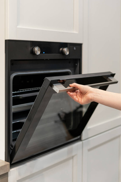 Foto vertical e real da porta de abertura da mão da mulher do novo forno preto embutido no armário da cozinha branca
 - Foto, Imagem