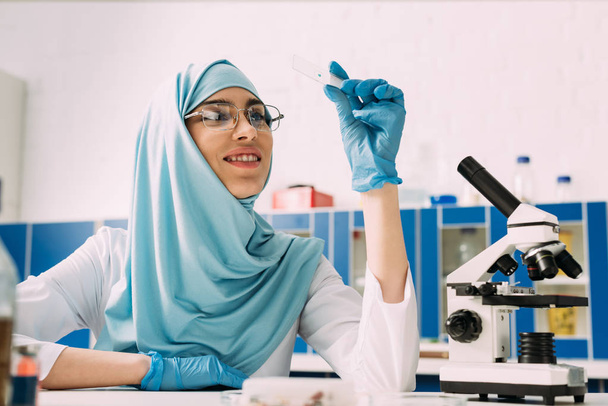 smiling female muslim scientist sitting at table with microscope and looking at glass sample during experiment in chemical laboratory - Foto, Imagen