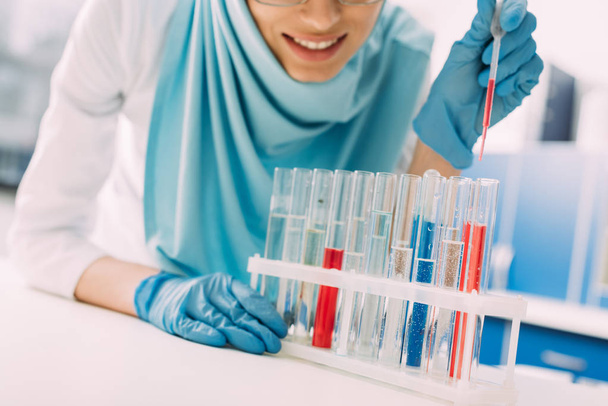 vista recortada de la sonriente científica musulmana que añade sangre en el tubo de ensayo en el laboratorio
 - Foto, Imagen