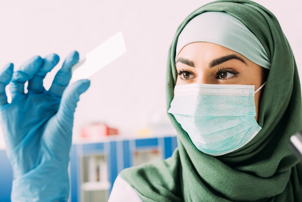 female muslim scientist in medical mask and hijab holding glass sample during experiment in laboratory - Фото, изображение