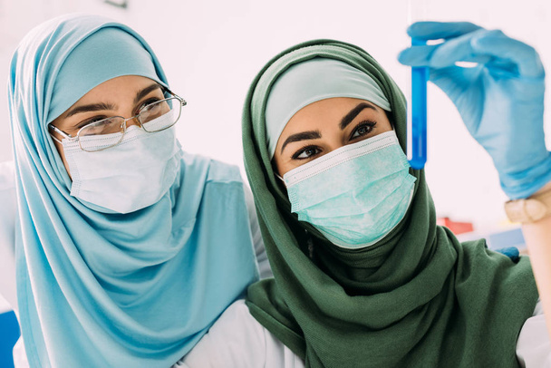 female muslim scientists in medical masks looking at glass test tube with reagent during experiment in chemical lab - Photo, Image