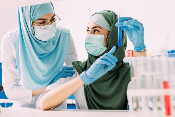 científicas musulmanas en máscaras médicas sosteniendo tubo de ensayo de vidrio con reactivo durante el experimento en laboratorio químico
 - Foto, Imagen