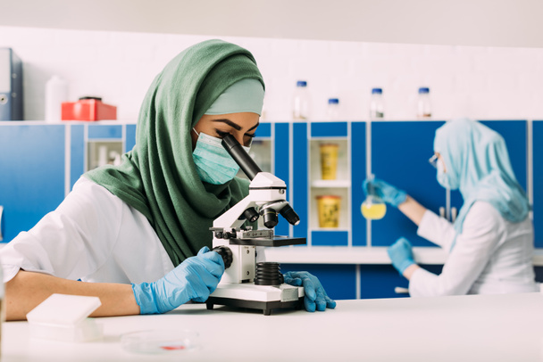 γυναίκα μουσουλμάνος επιστήμονας αναζητούν μέσα από το μικροσκόπιο κατά τη διάρκεια πειράματος με συνάδελφο που εργάζεται στο υπόβαθρο σε χημικό εργαστήριο - Φωτογραφία, εικόνα