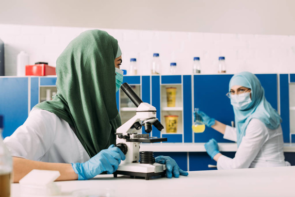 γυναίκα μουσουλμάνος επιστήμονας χρησιμοποιώντας μικροσκόπιο κατά τη διάρκεια πειράματος με συνάδελφο που εργάζεται στο υπόβαθρο σε χημικό εργαστήριο - Φωτογραφία, εικόνα