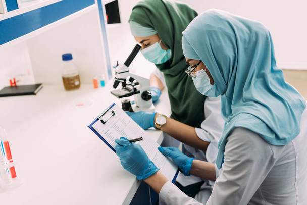 Muslimische Wissenschaftlerinnen in medizinischen Masken mit Mikroskop und Klemmbrett während eines Experiments im Chemielabor - Foto, Bild