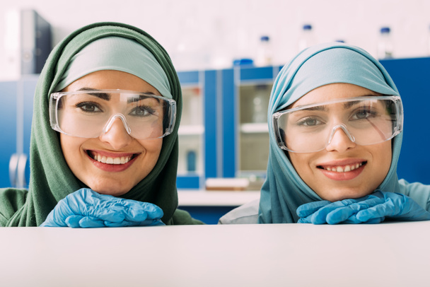 smiling female muslim chemists in protective goggles and hijab looking at camera in laboratory - Foto, Imagem