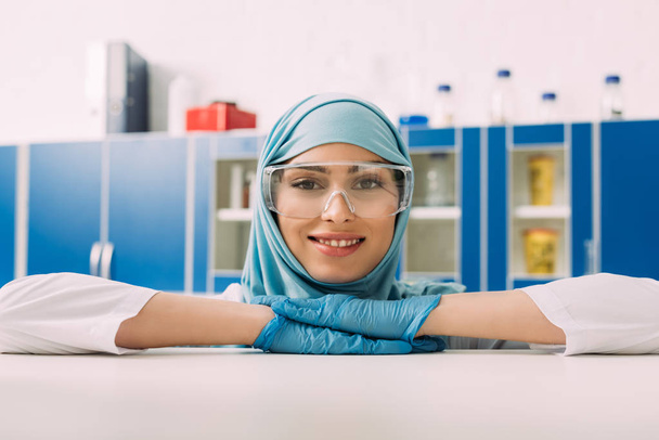 ラテックス手袋、ゴーグル、ヒジャーブの研究室ではカメラ目線で笑顔の女性イスラム教徒の科学 - 写真・画像