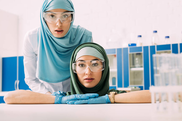 female muslim chemists in protective goggles and hijab looking at camera in laboratory - Photo, Image