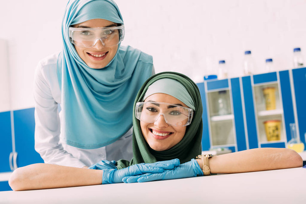 ευτυχισμένος μουσουλμανική χημικοί προστατευτικά γυαλιά και χιτζάμπ βλέπουν φωτογραφική μηχανή στο εργαστήριο - Φωτογραφία, εικόνα