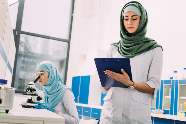 concentrated female muslim scientists using microscope and writing in clipboard during experiment in chemical laboratory - Photo, Image