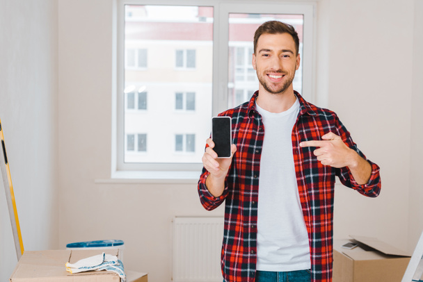 cheerful man pointing with finger at smartphone with blank screen while standing at home - Foto, Imagem
