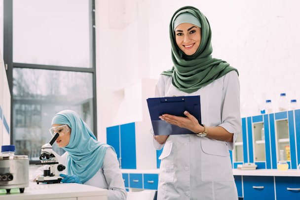 θηλυκό μουσουλμάνων επιστημόνων που χρησιμοποιώντας το μικροσκόπιο και πρόχειρο κατά τη διάρκεια του πειράματος σε χημικό εργαστήριο - Φωτογραφία, εικόνα