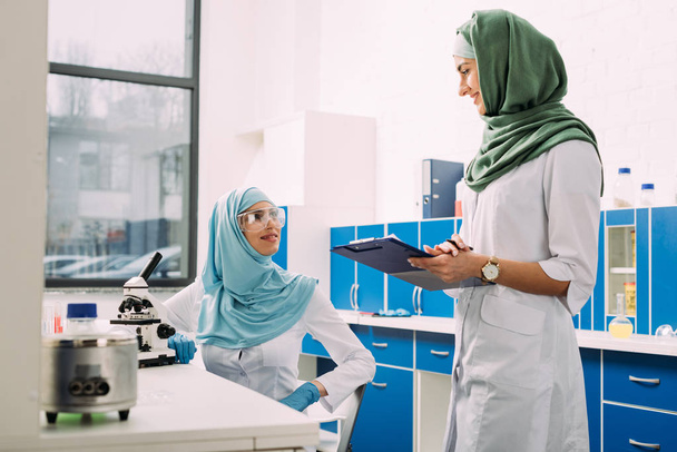 θηλυκό μουσουλμάνων επιστημόνων που χρησιμοποιώντας το μικροσκόπιο και πρόχειρο κατά τη διάρκεια του πειράματος σε χημικό εργαστήριο - Φωτογραφία, εικόνα