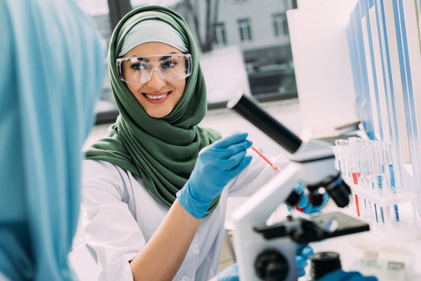 Lachende vrouw moslim wetenschapper in bril en hijab tijdens experiment in chemisch laboratorium - Foto, afbeelding