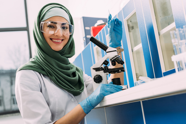 женщина-мусульманский ученый сидит за столом с микроскопом, держит образец стекла и смотрит в камеру в химической лаборатории
 - Фото, изображение