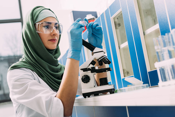 γυναίκα μουσουλμάνος επιστήμονας κάθεται στο τραπέζι με μικροσκόπιο και κοιτάζοντας γυαλί δείγμα κατά τη διάρκεια του πειράματος σε χημικό εργαστήριο - Φωτογραφία, εικόνα