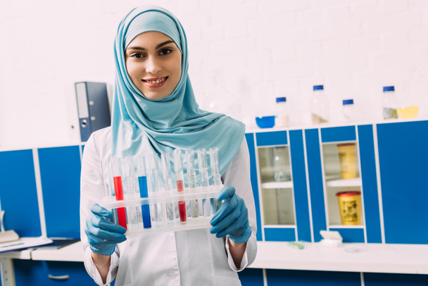 sonriente mujer musulmana científica sosteniendo tubos de ensayo con líquido rojo y azul mientras mira a la cámara
 - Foto, Imagen