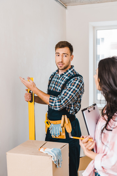 selective focus of handyman gesturing and holding measuring level near woman with clipboard and looking at wall  - Foto, afbeelding